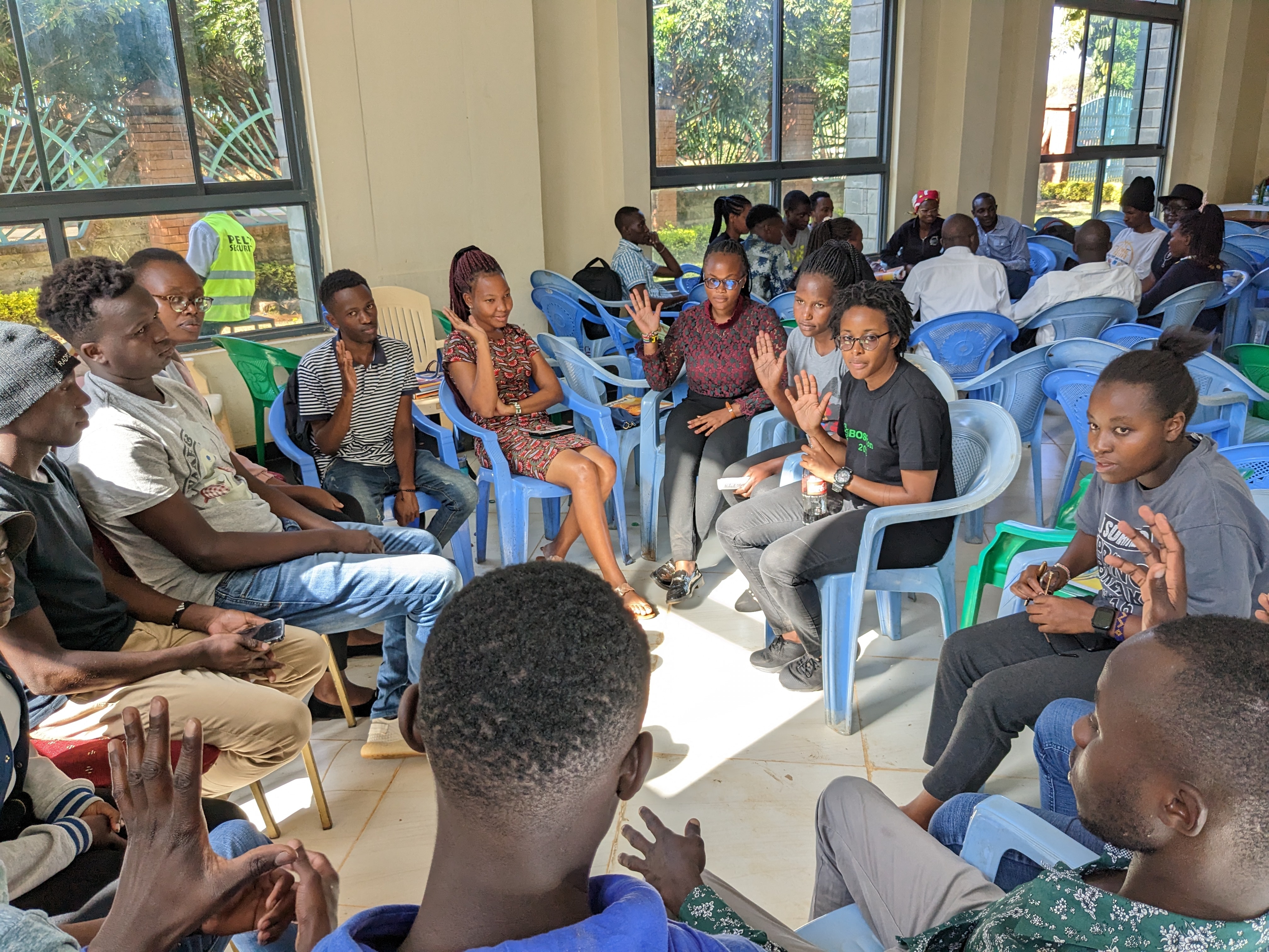 Integrantes del Bioinformatics Hub of Kenya, jóvenes estudiantes, aproximadamente 6 mujeres y cinco hombres, sentados en sillas azules formando un círculo, dentro de un moderno edificio de hormigón con techos altos, muchos de ellos levantando la mano como en una votación, entablando una conversación, la luz del sol crea una forma cuadrada en el centro del círculo